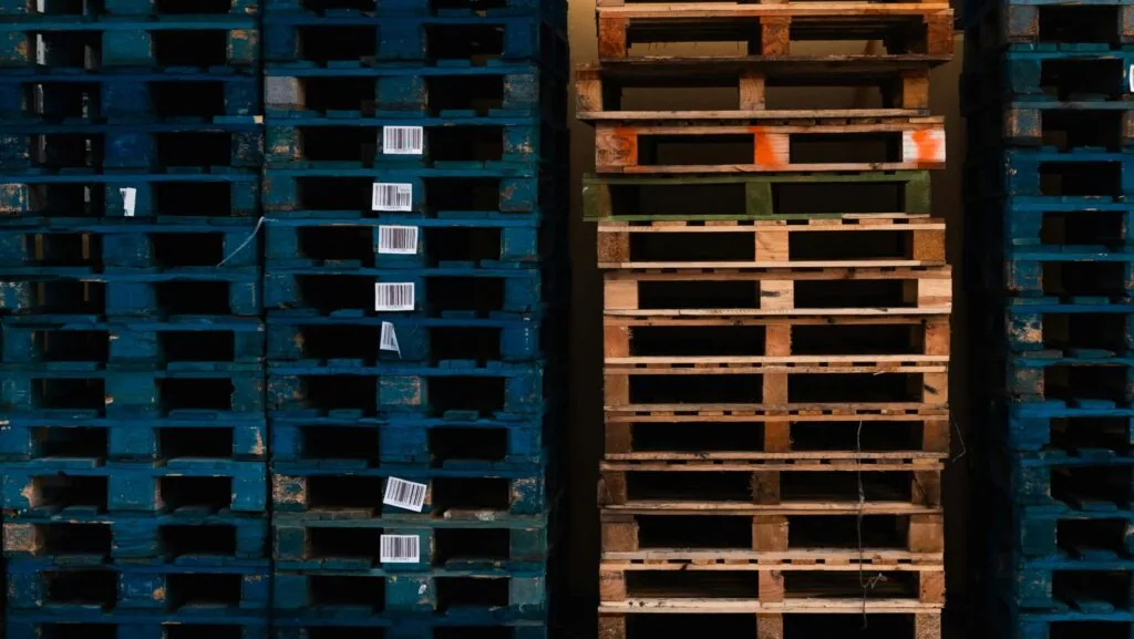 Stacks of blue and brown wooden pallets, including some custom-sized pallets, are arranged next to each other; the blue pallets have barcode stickers attached to some slats.